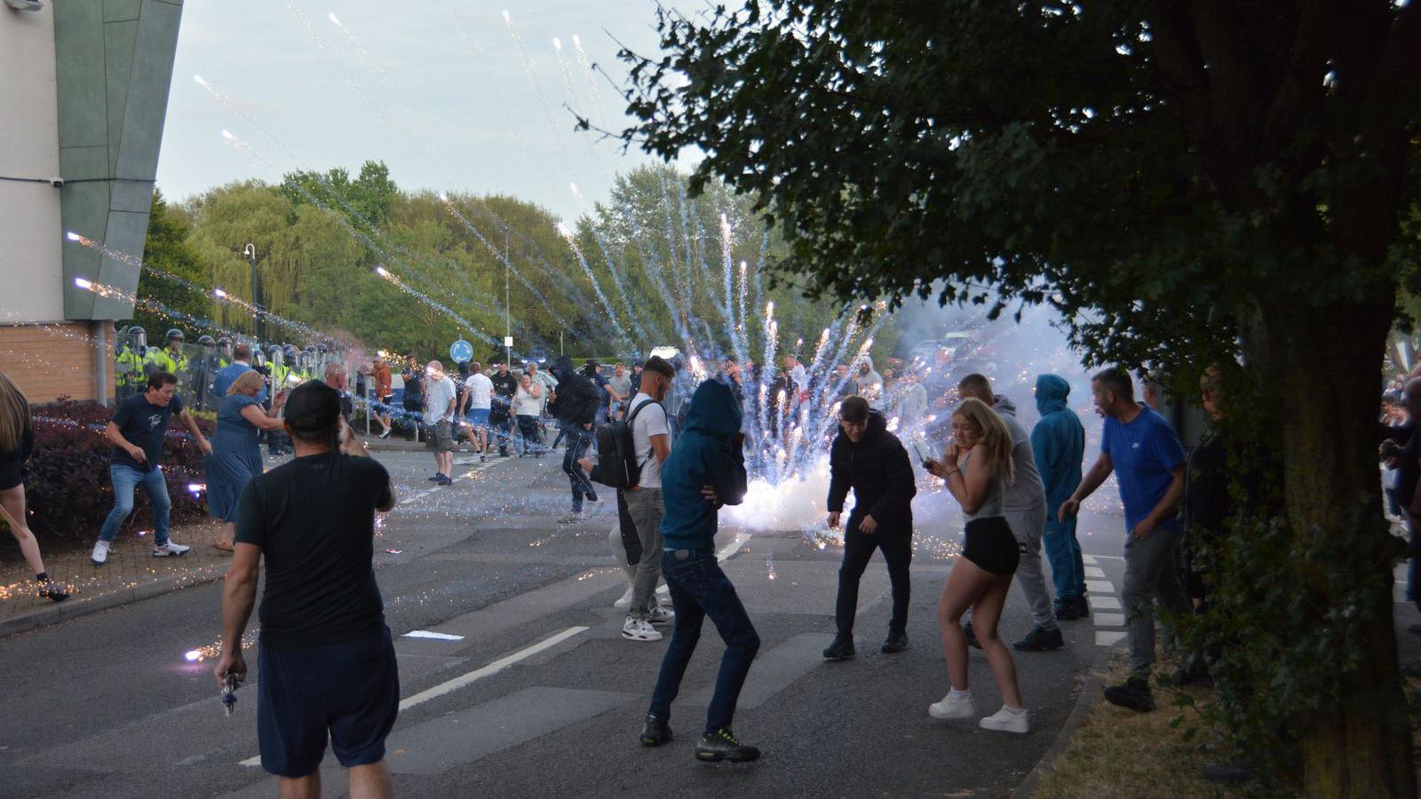 A group of people stand on a street as a firework explodes in the middle of a road outside a hotel building. Several are turning away amid the explosion.  