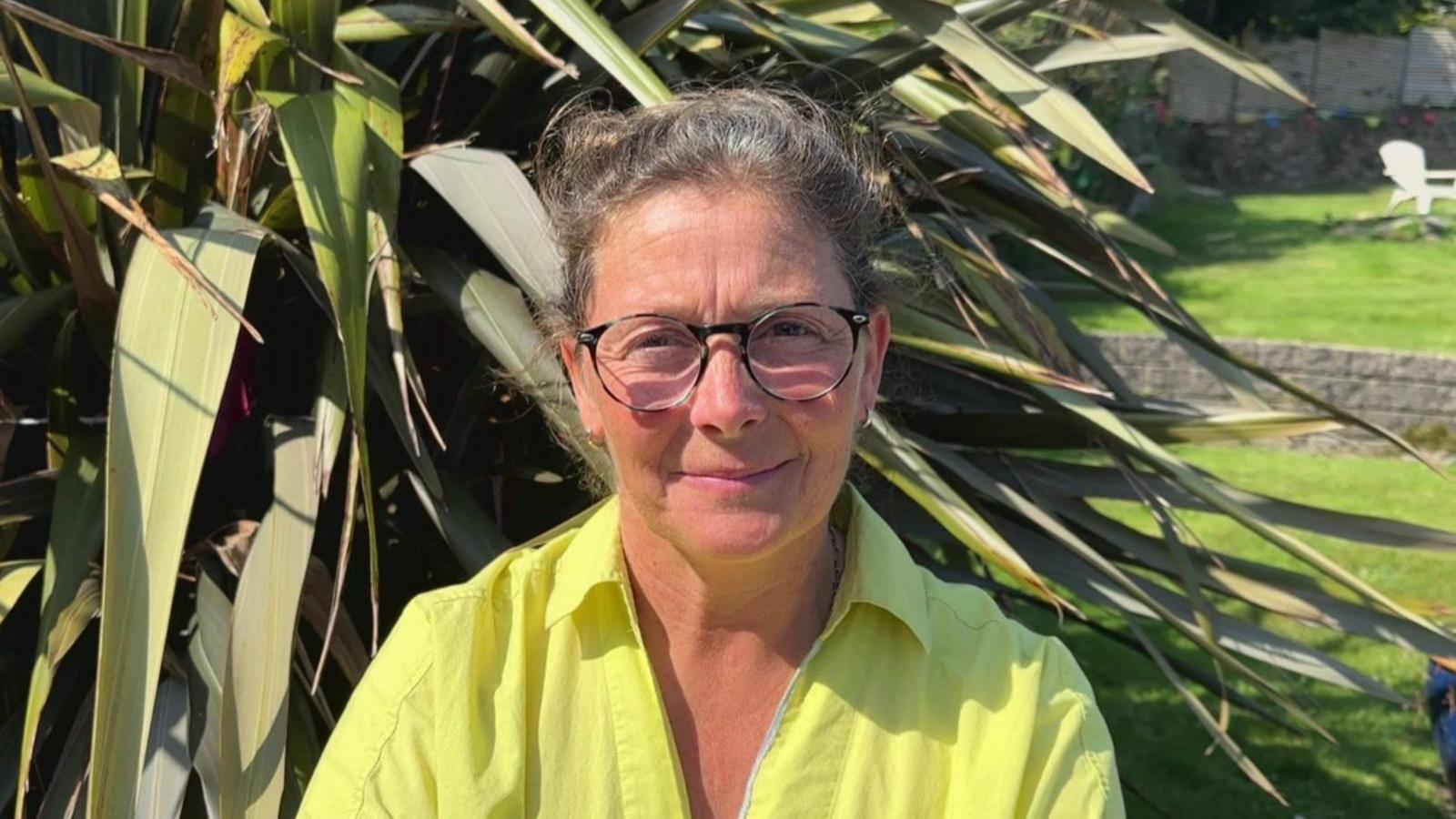 Claire Bailey, a doctor at the Royal Cornwall Hospital. She is pictured in front of a plant. She is wearing a florescent green shirt and has black glasses. 