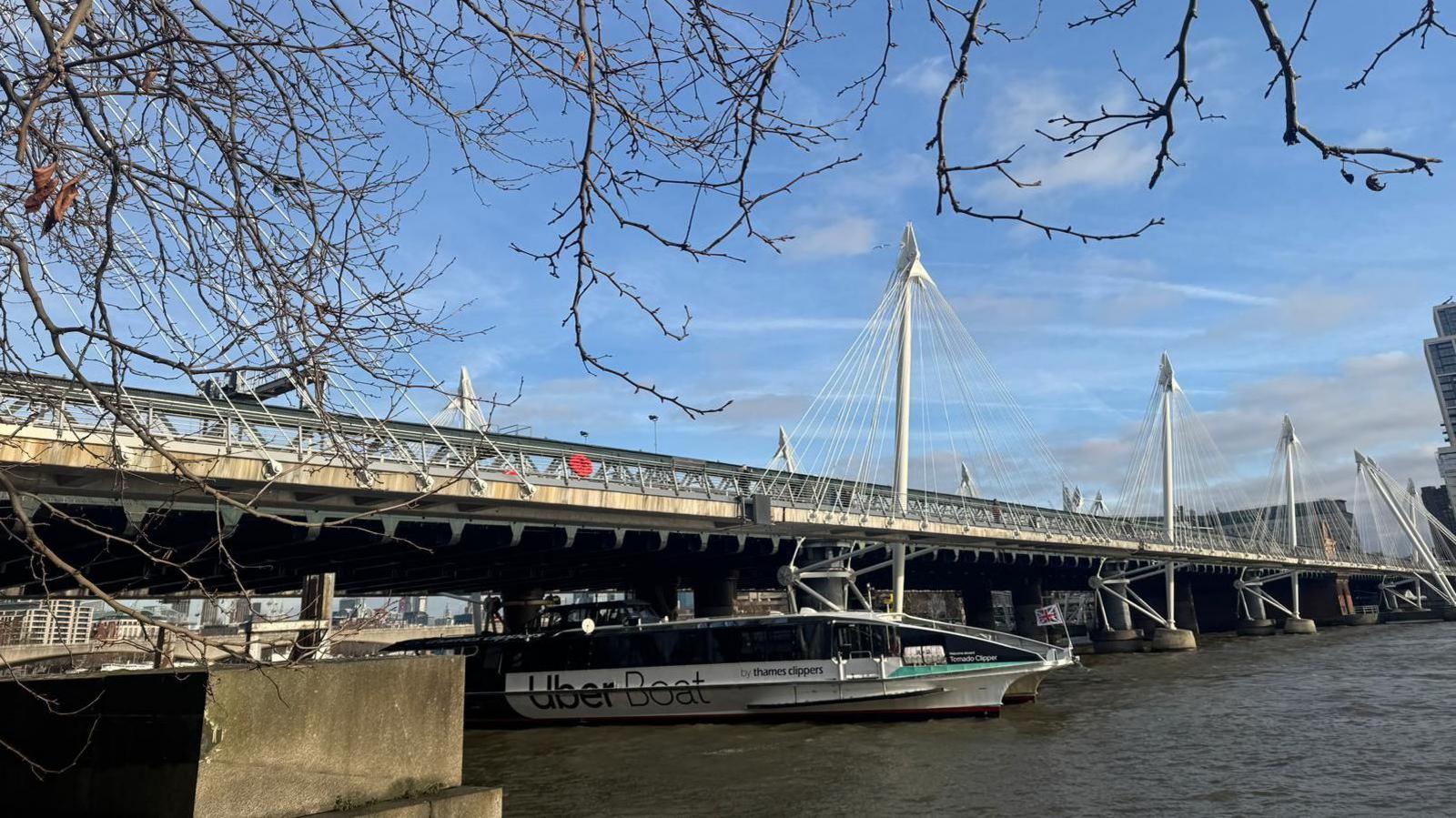 A landscape shot of the Victorian-style bridge from Victoria embankment with the River Thames running beneath it and bright blue sky. Outward leaning pylons run along each side and there is an Uber Boat in motion beneath the bridge. 