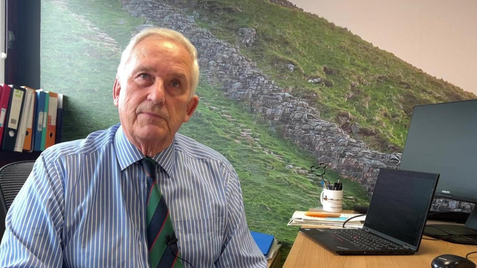 Glen Sanderson sits at his desk, wearing a blue and white striped shirt with a green and navy blue tie. A vinyl print of Hadrian's Wall covers the wall behind him while on the desk sits a computer and a mug holding various pens. 