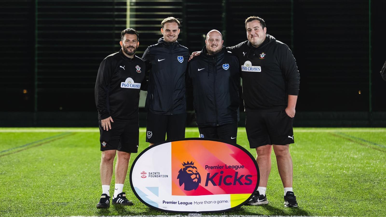 Four men, two wearing Southampton football club tracksuit kits (from the Saints Foundation) and two wearing Portsmouth Football club tracksuit kits (From Pompey in the Community). They are stood in a row with their arms around each other on a green astro turf pitch with a sign in front of them which reads "Premier League Kicks" 
