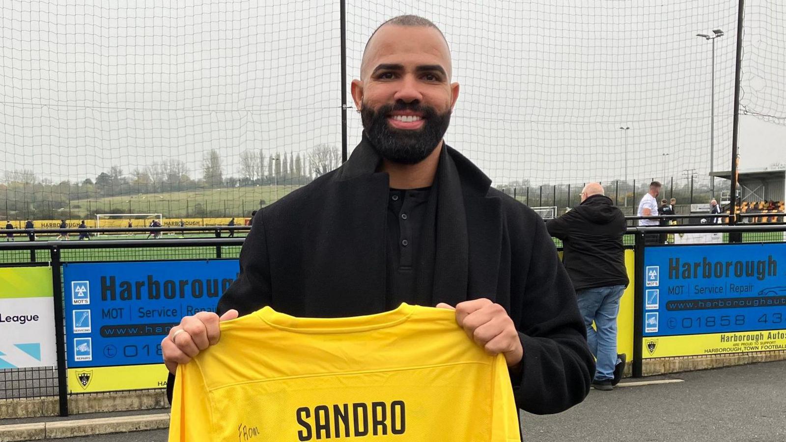 Former Tottenham and Brazil midfielder Sandro holds up a Harborough Town shirt as he watches the non-league club he has just signed for