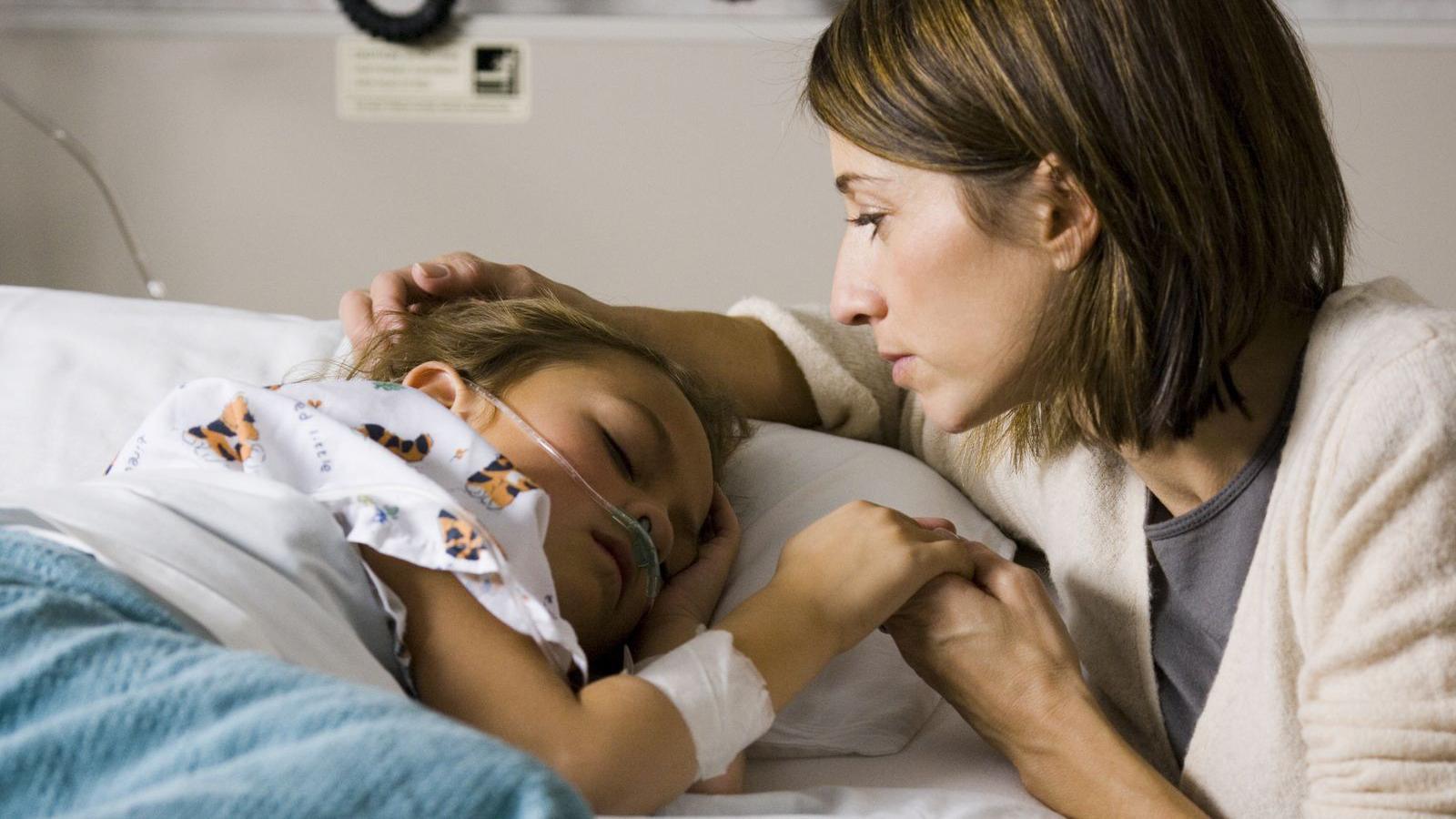 A child laying down in a hospital bed with brown hair and a tube attached, she is wearing white clothing with animals on it and is lying under a blanket. A woman is holding her hand and has her hand on the child's head, she has short brown hair and is wearing a cream cardigan with a grey top.