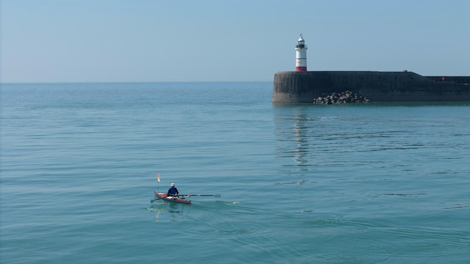 Mr Ellicock in his boat and heading out to sea. The water is still and blue and the sky is also clear and blue. In the background there is a lighthouse on a harbour wall.