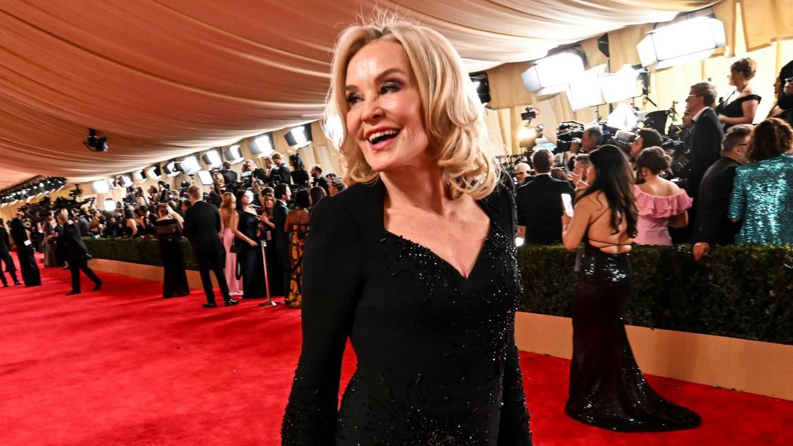 Jessica Lange - a woman with blond hair and in a black dress - smiles and laughs while on the red carpet at the 2024 Oscars