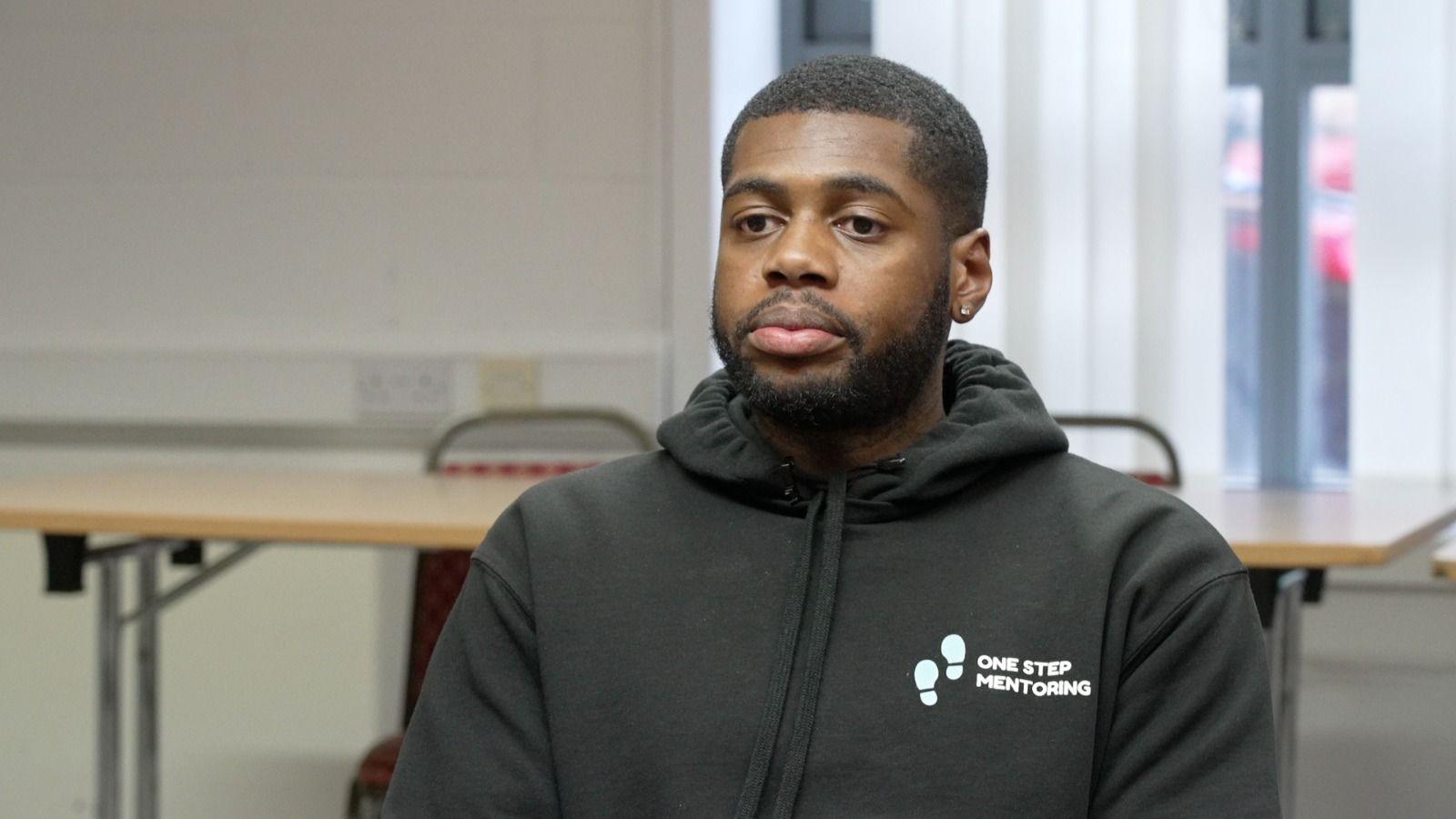 Alex McLean sits in a room infront of tables and chairs, he is wearing a black hooded top with a logo 'One Step Mentoring' 