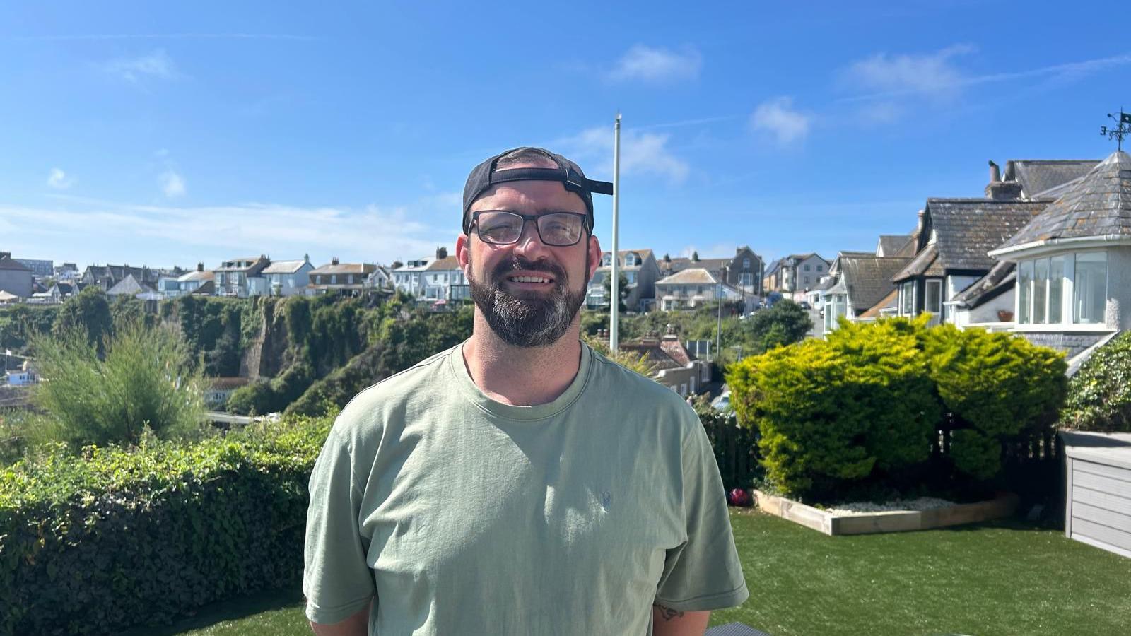 Matt, Anya's dad, is stood in a garden with houses and other gardens behind him and houses to the right. He is wearing a light green top, glasses and a baseball cap on backwards.