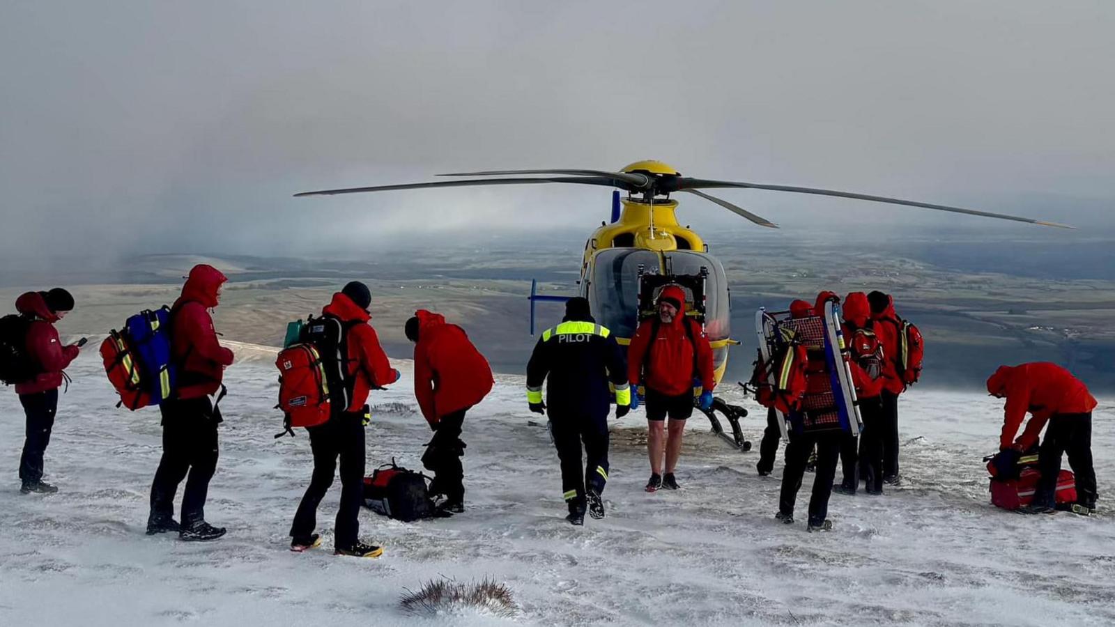 A team of Keswick Mountain Rescue volunteers, who are all wearing red waterproofs and large rucksacks, stand on top of the summit of Blencathra. There is a yellow helicopter behind them.