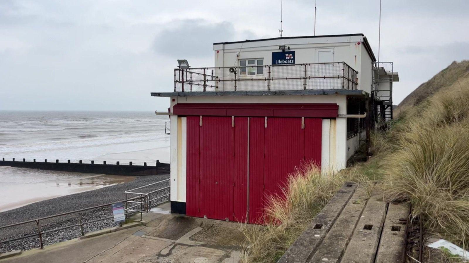 View of the Sheringham lifeboat shed. A building on the cliffside with red doors overlooking the pebble beach and sea beyond. 