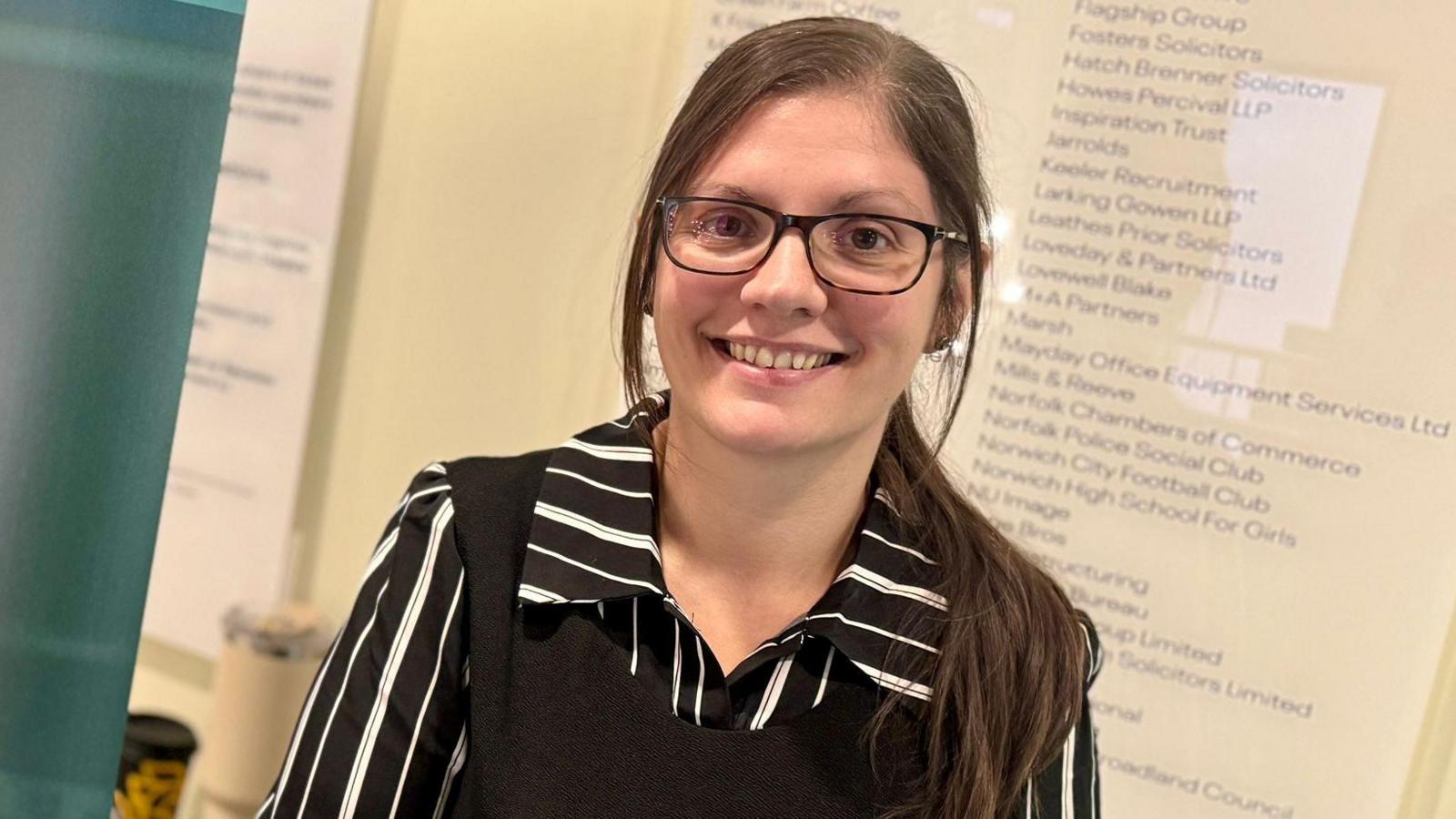 Charlotte Notley smiles at the camera. She is wearing a black and white striped shirt with a black jumper on top. She has dark glasses and is standing in front of a board with names on it.