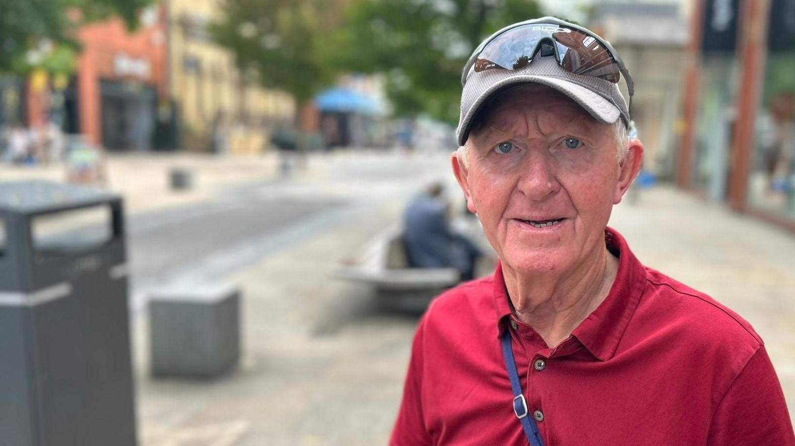 Colin Richardson, with white hair, wears a grey baseball cap with sunglasses on top. He is sporting a red open-necked shirt.