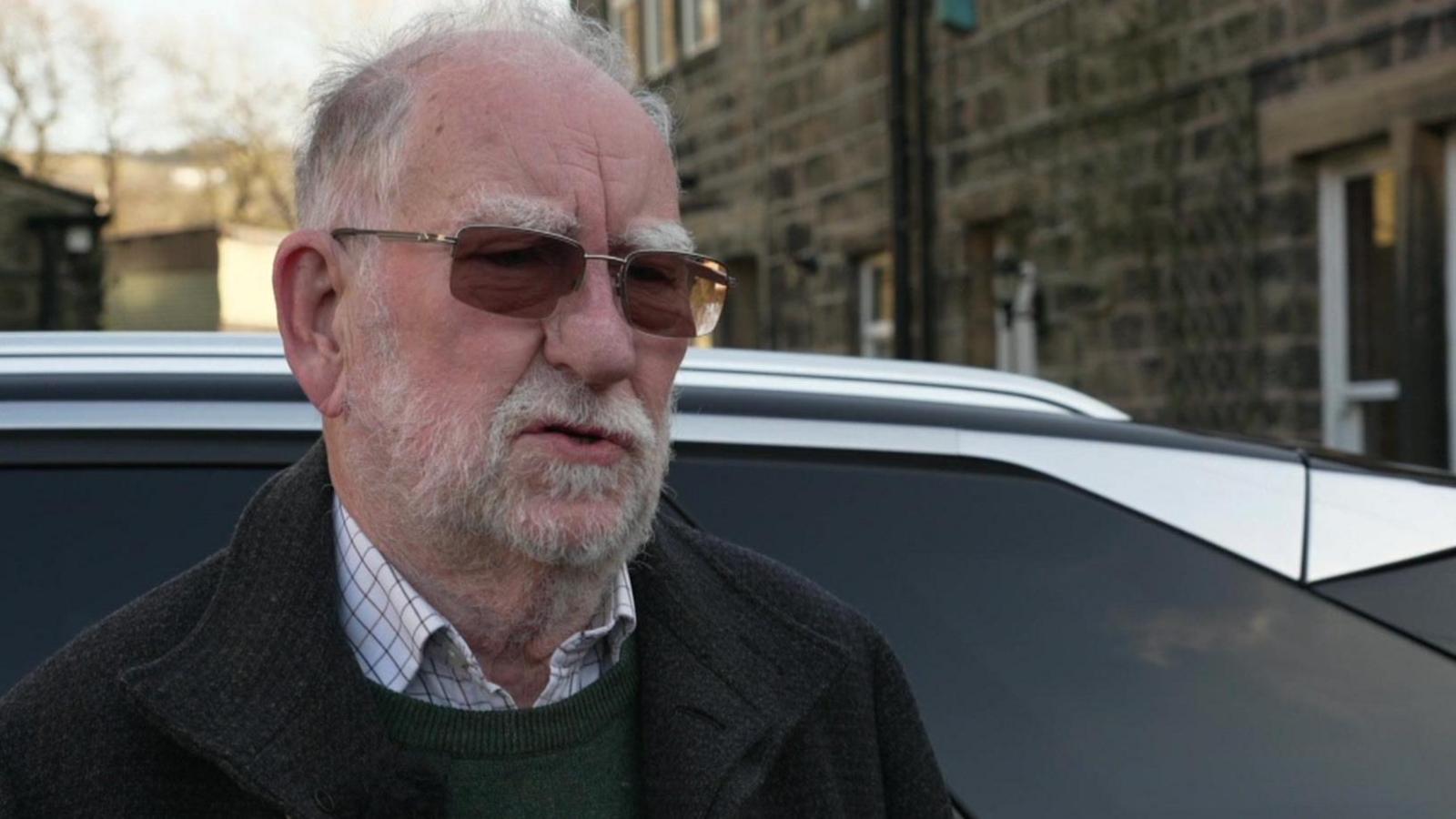 An older man with a white beard and wearing dark lens glasses stands in front of a car, itself next to what looks like a cottage built in Yorkshire stone.