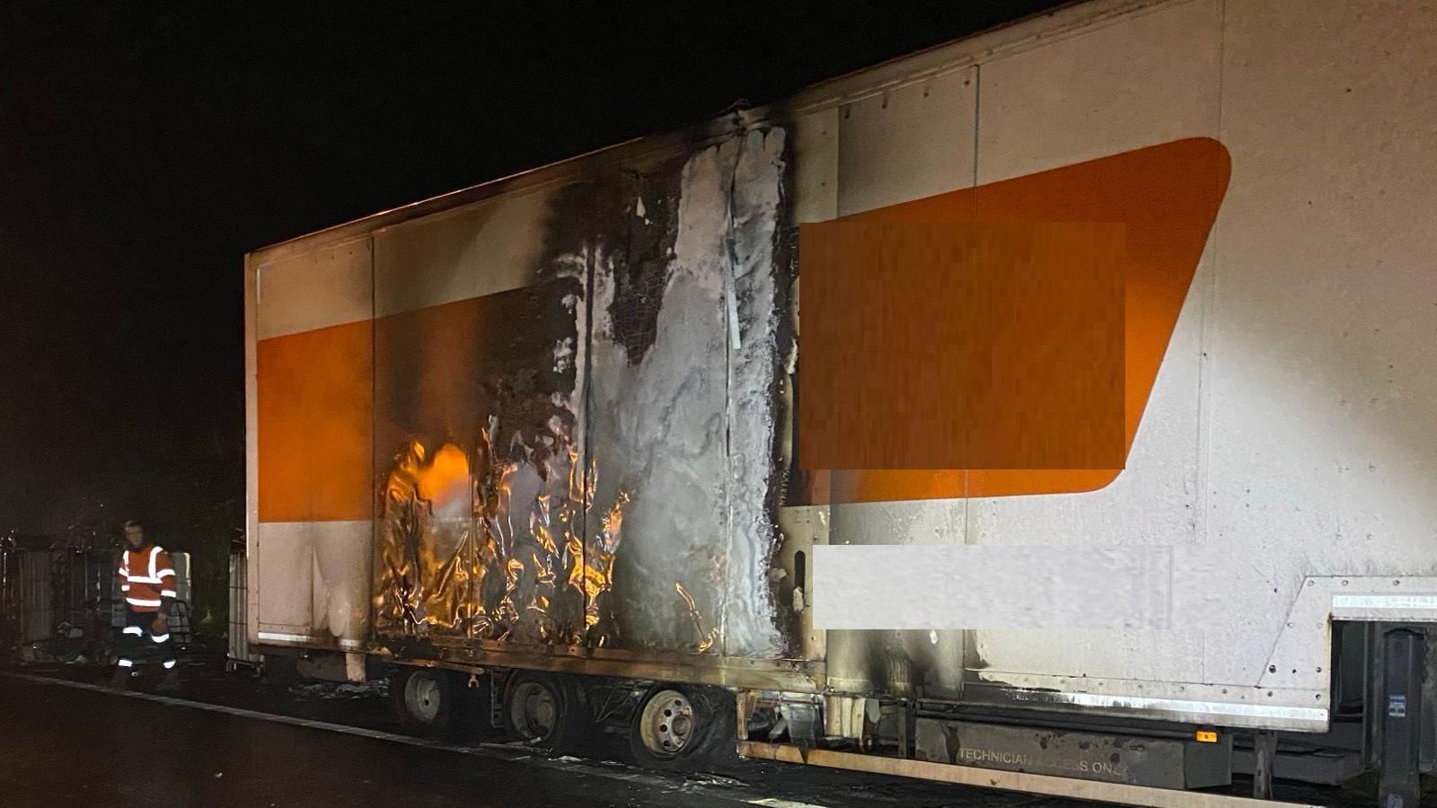 A lorry on fire on the M45 in the dark. The vehicle has a white and orange side, with flames coming out it and visible damage. A man in a high vis outfit is standing at the rear end of the trailer. 