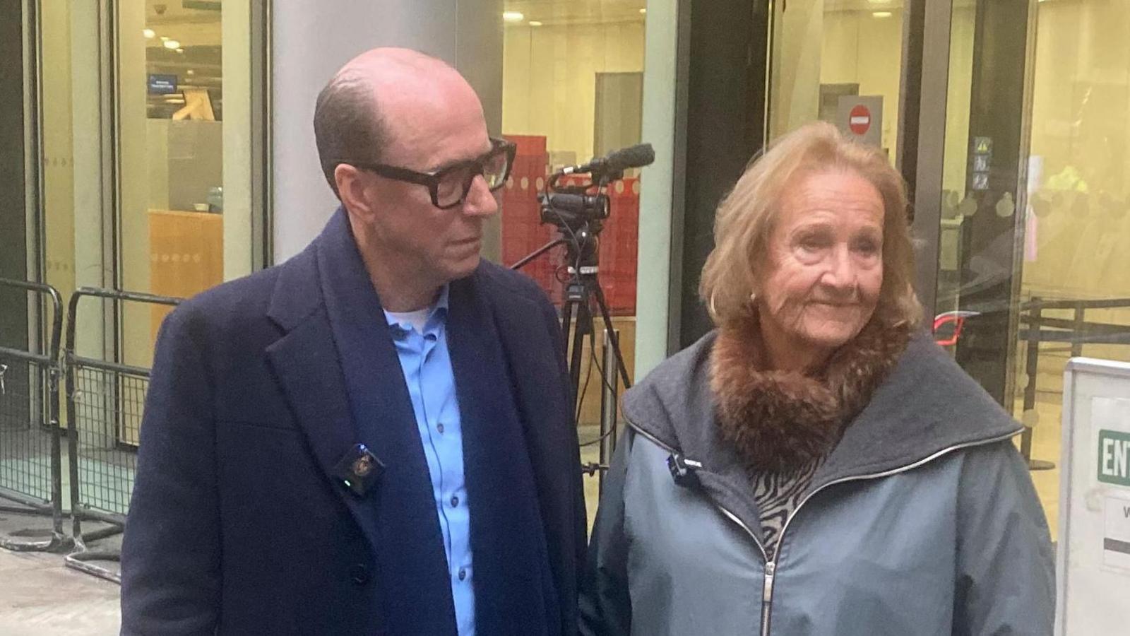 A man and a woman standing on a paved walkway outside a modern glass-fronted building. Both are dressed in coats suitable for cool weather; one wears a dark overcoat with a scarf, while the other has a light grey coat featuring a fur-trimmed collar. Behind them, there is a tripod with a mounted camera, suggesting media coverage or filming.