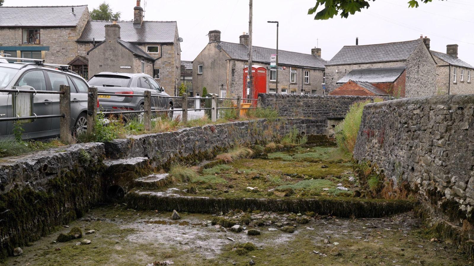 The bed of the brook is covered only in stones and mosses rather than filled with water. Either side is stone walls. On the left-hand side are two grey parked cars, a red telephone box and a few houses.
