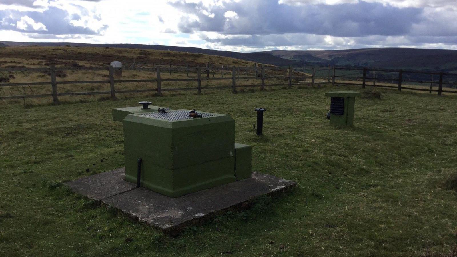 Small green-painted square structures and a metal pipe sticking out of the ground surrounded by a fence and in a hilly landscape.