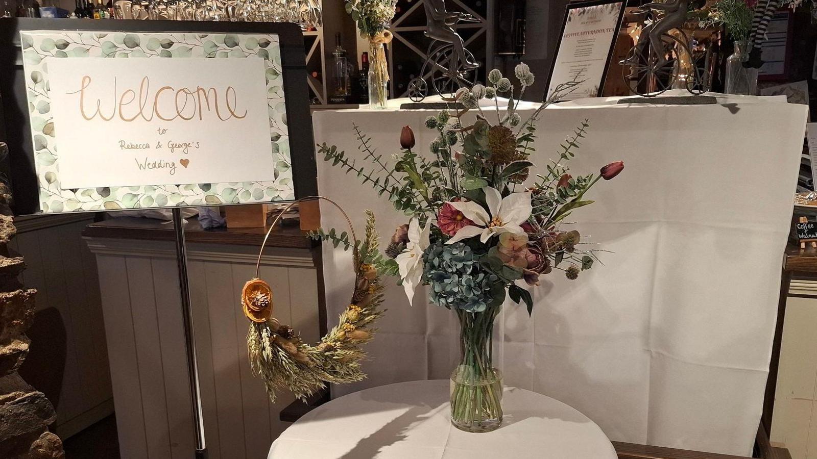 Welcome sign at entrance to restaurant with the couple's names and a round table with white table cloth and bunch of flowers in a glass vase. A wreath made of flowers and grasses hangs from the welcome sign 