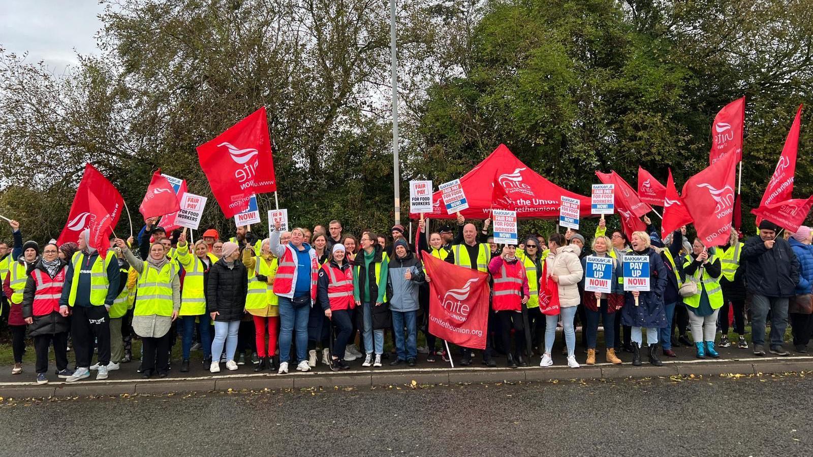 A large group of factory workers standing on a path outside wearing yellow or red hi-vis jackets. Many are holding red banners or flags branded with Unite the Union's logo. Others are holding banners that read 'pay up now!' There are trees behind them with green leaves and the sky is cloudy. 