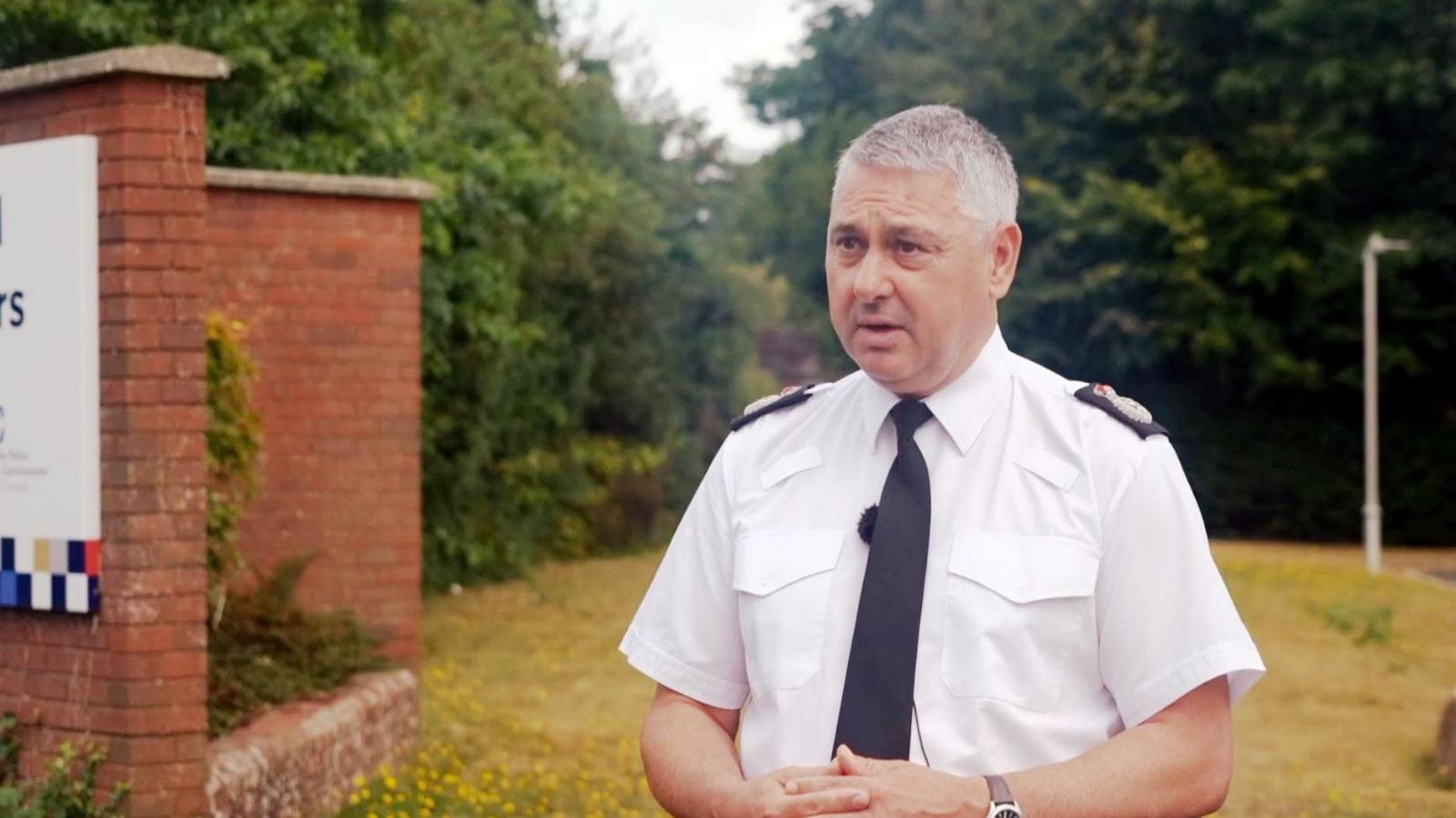A grey haired man in a short-sleeved white police shirt with a black tie. He stands in front of some trees and a patch of grass, next to the a sign.