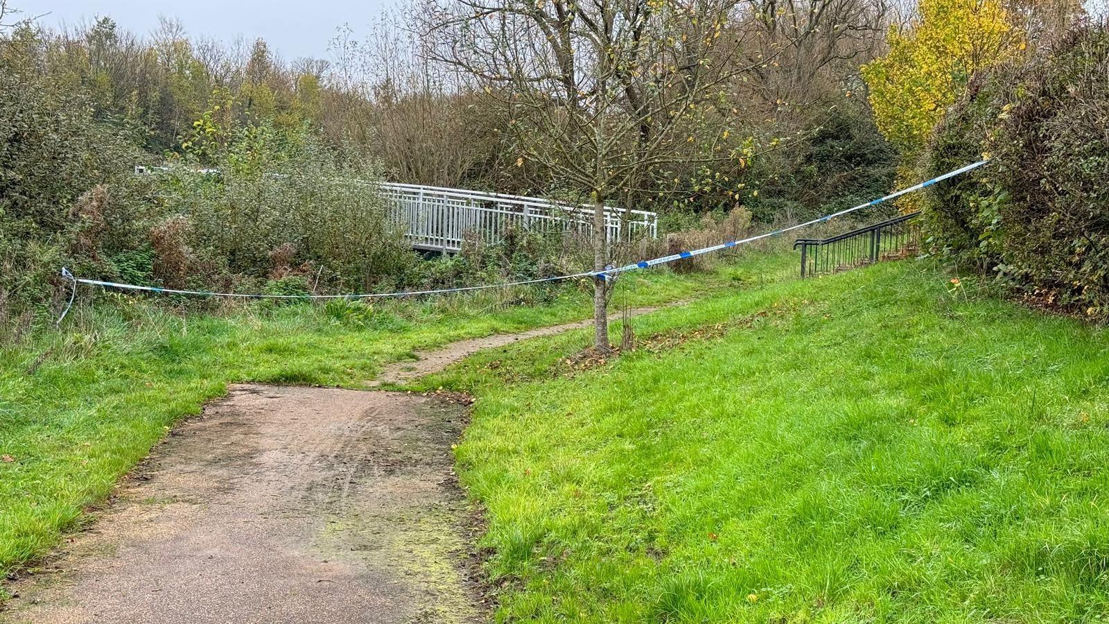 Police tape across a muddy path. There are bushes on either side and a metal bridge can be seen to the left. 