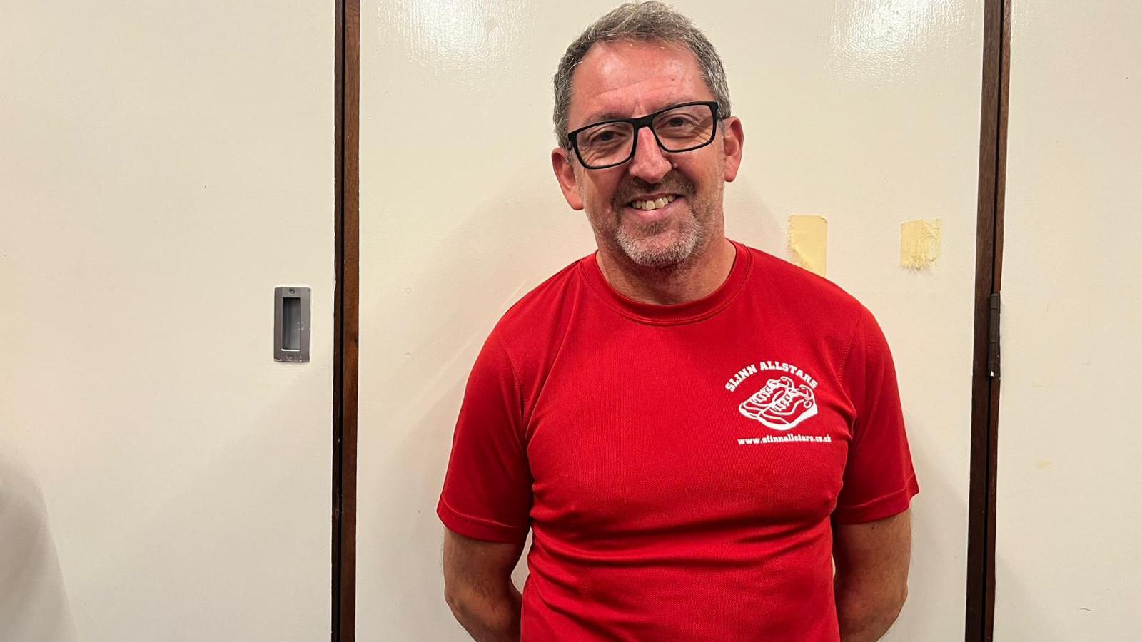 One man standing against a cream-coloured wall with wooden trim. The man is wearing a plain bright red T-shirt featuring the “Slinn Allstars Swindon” logo and website text on the chest. The background is simple, with a small metal fixture visible on the wall.