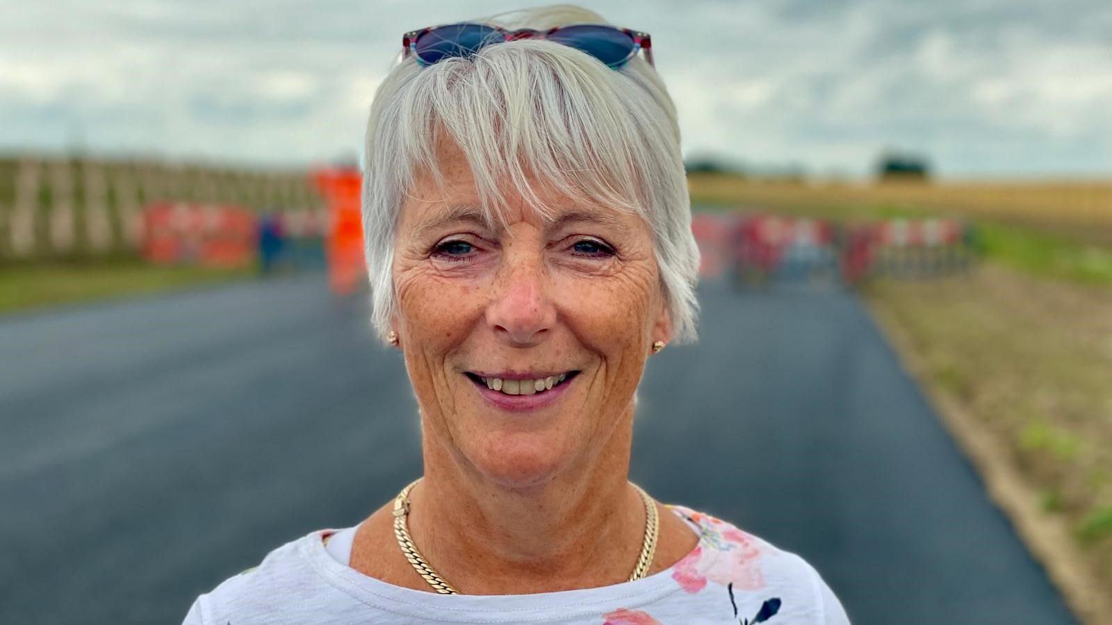 A woman with white hair smiles as she faces the camera, with a road, grass and safety barriers seen behind her out of focus.