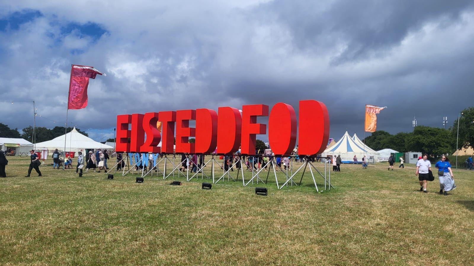 A large letter display reading "Eisteddfod" can be seen in red lettering in the centre of a field at the Welsh national festival. Large white tents can be seen in the background along with people walking around the site.
