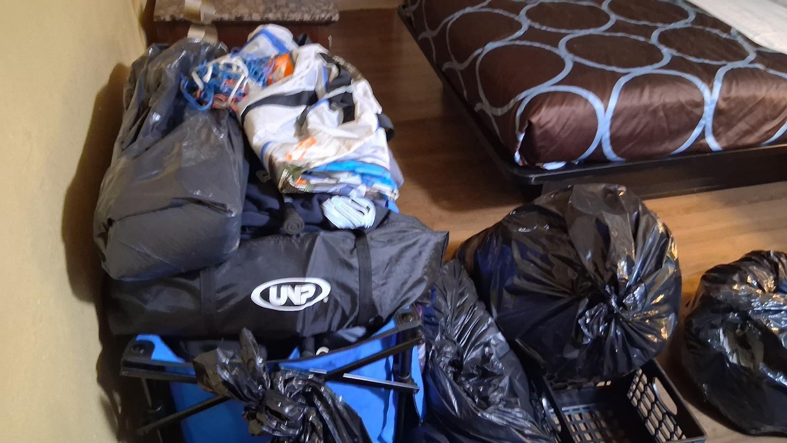 Duffel bags and full garbage bags are piled up on a floor next to a bed with a brown duvet