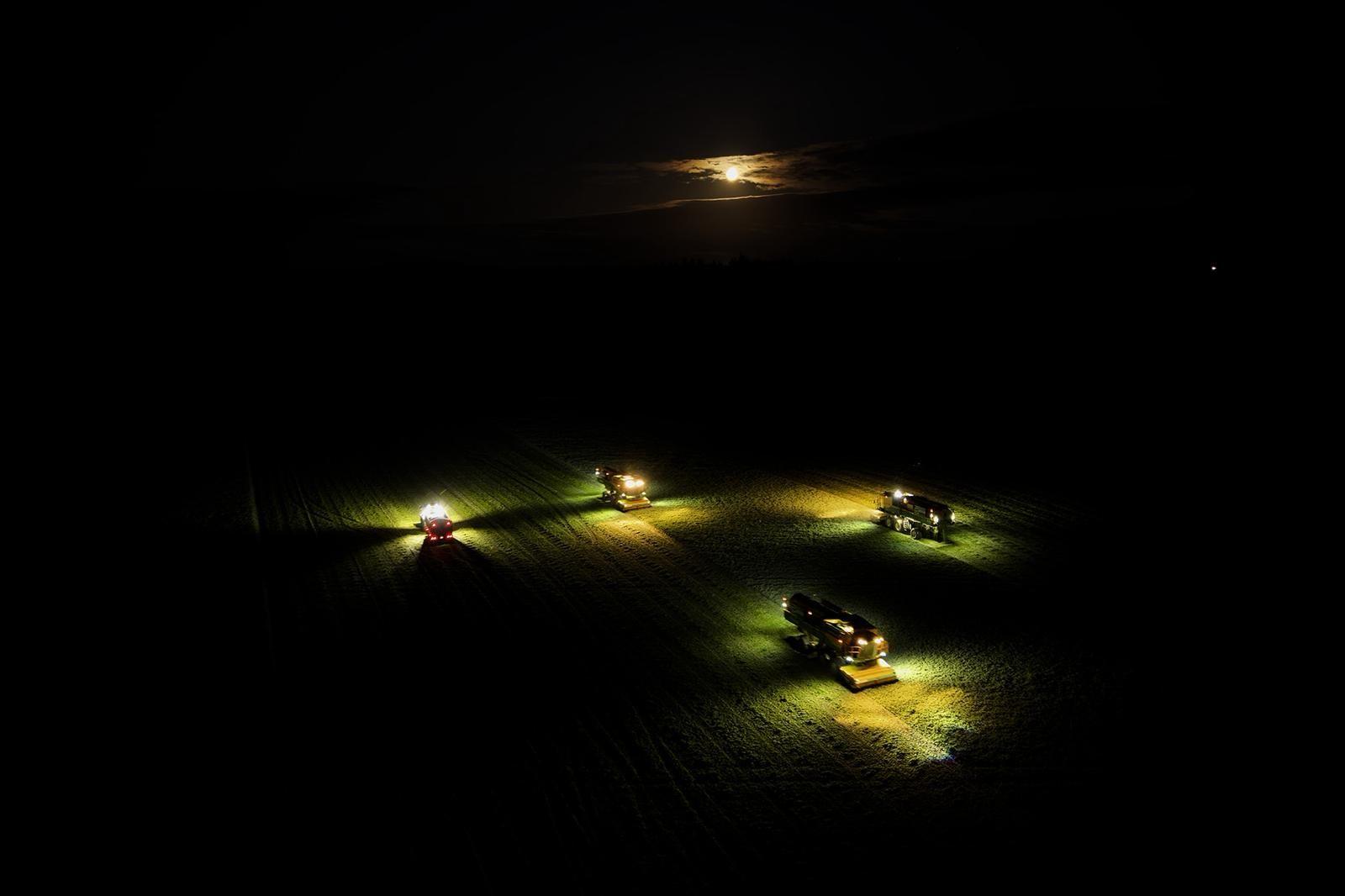 Aerial image of harvesting machinery with bright lights shining on a field in pitch black darkness with only the moon peaking above the clouds.