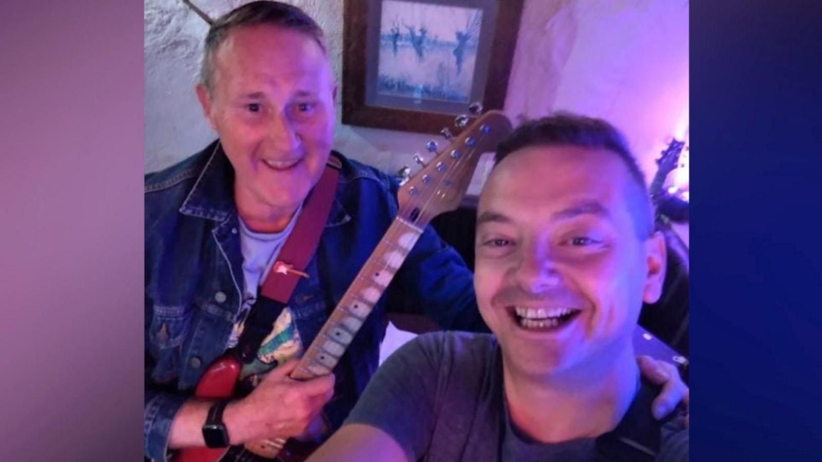 Dean and Tony smile at the camera as Dean takes a selfie. Dean is wearing a grey t-shirt and has short dark hair. Tony is wearing a denim jacket and has a red guitar with one hand and has his other hand on Dean 's shoulder. They're in purple lighting.