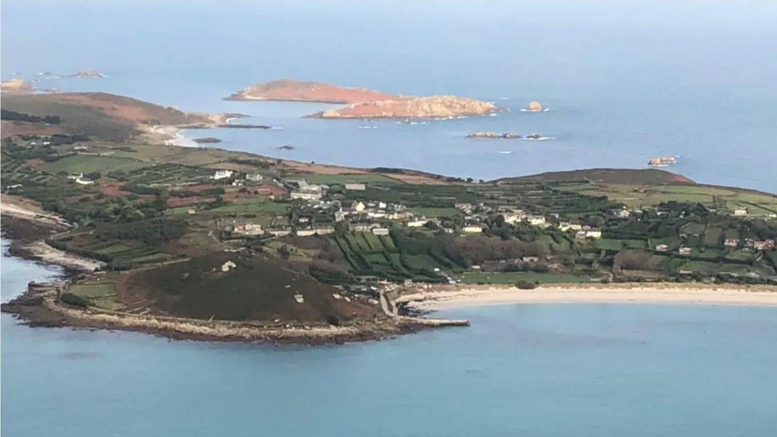 An aerial view of Isles of Sciily showing a crescent-shaped beach, green countryside and a small number of houses.