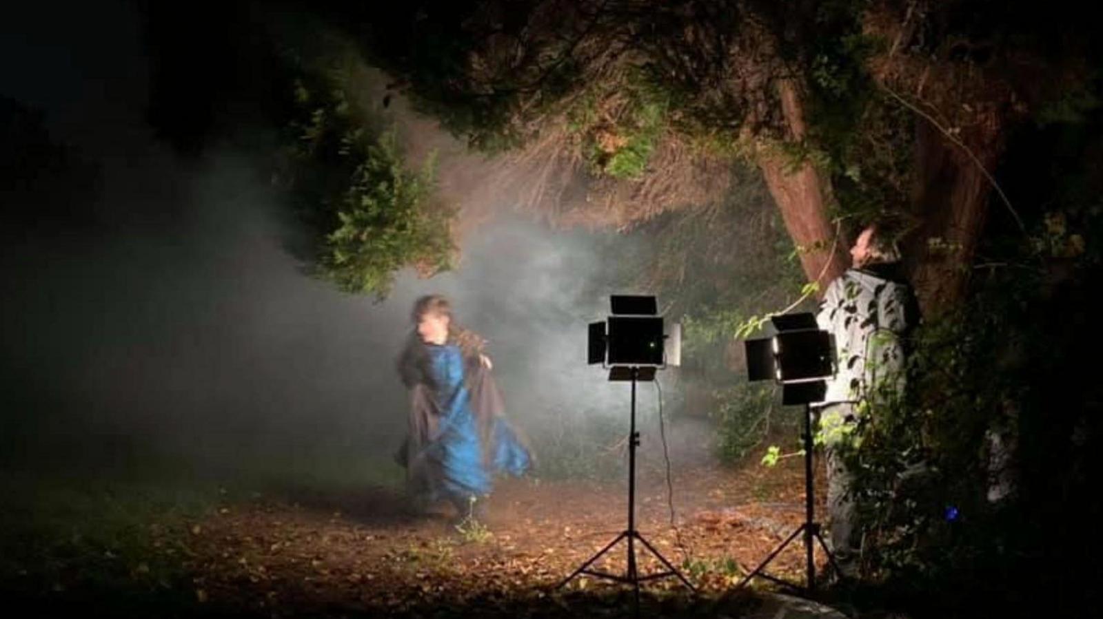 An actress in Victorian clothing running through a dark park. She is illuminated by stage lights. A member of the film crew stands behind the lights.