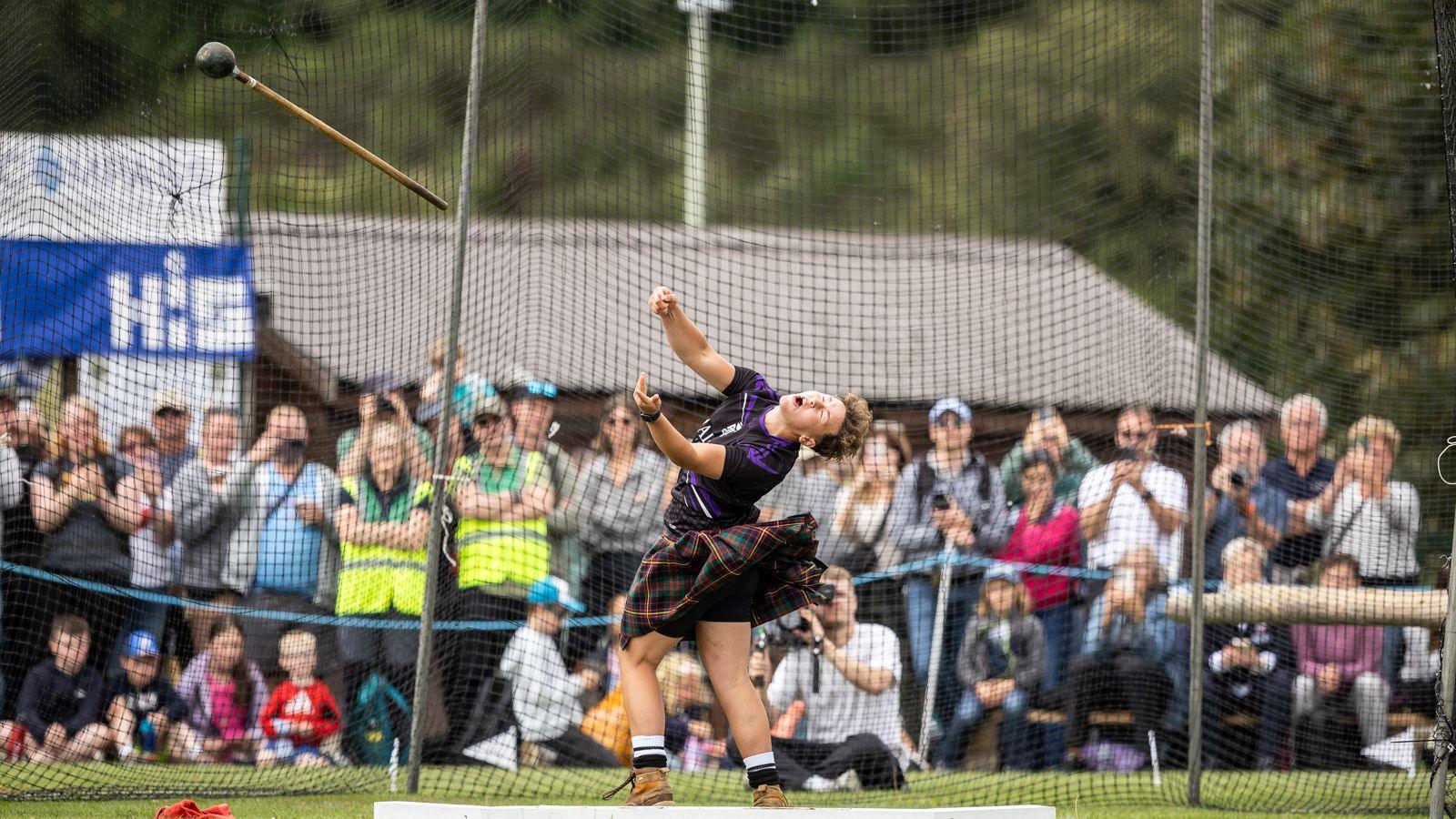 In an action shot, an athlete throws the Scots hammer. Her eyes are closed as she sends the hammer flying. Behind her is a large net to prevent the weight from accidentally being thrown into the crowd standing around the events arena.