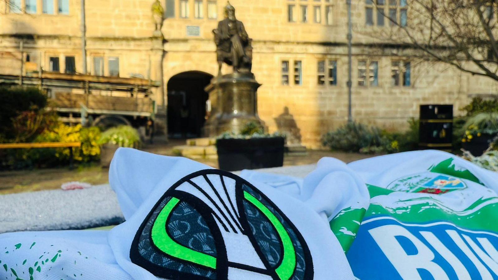 A football shirt on a wall, with Charles Darwin statue (a man sitting in a chair on a plinth) and Shrewsbury library (sandstone brick building) in the background. The football shirt is white with green stripes.