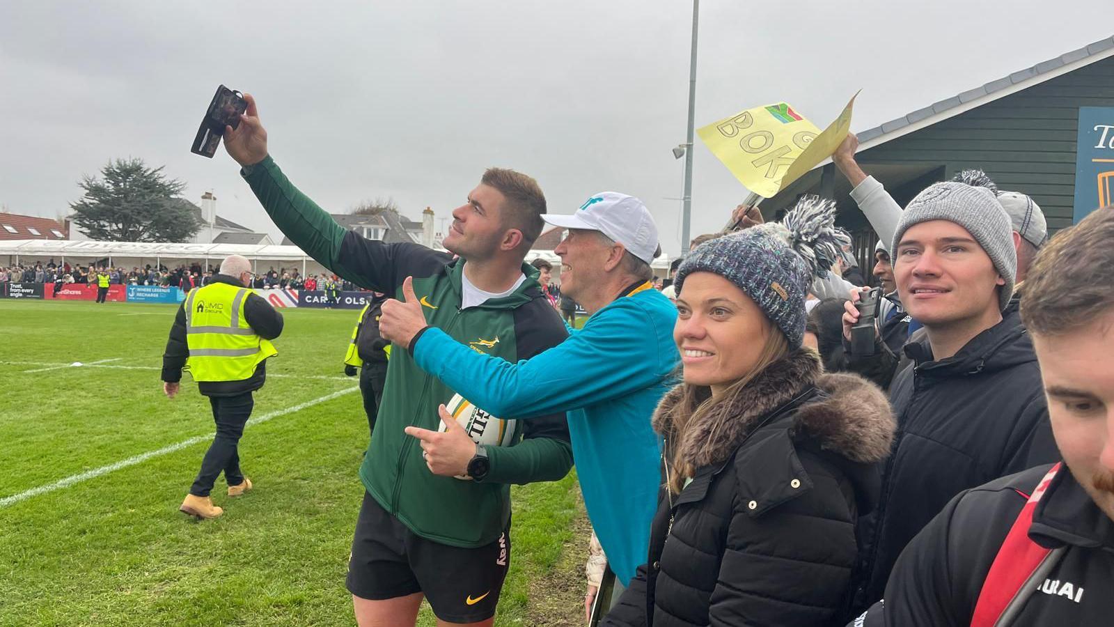 A Springbok player on the pitch holding a mobile phone in his right-hand taking a selfie with a fan. The man, who he is taking a selfie with, is holding up his thumb and smiling in the camera. He is wearing a blue T-shirt. On the right there are more people watching the training session.