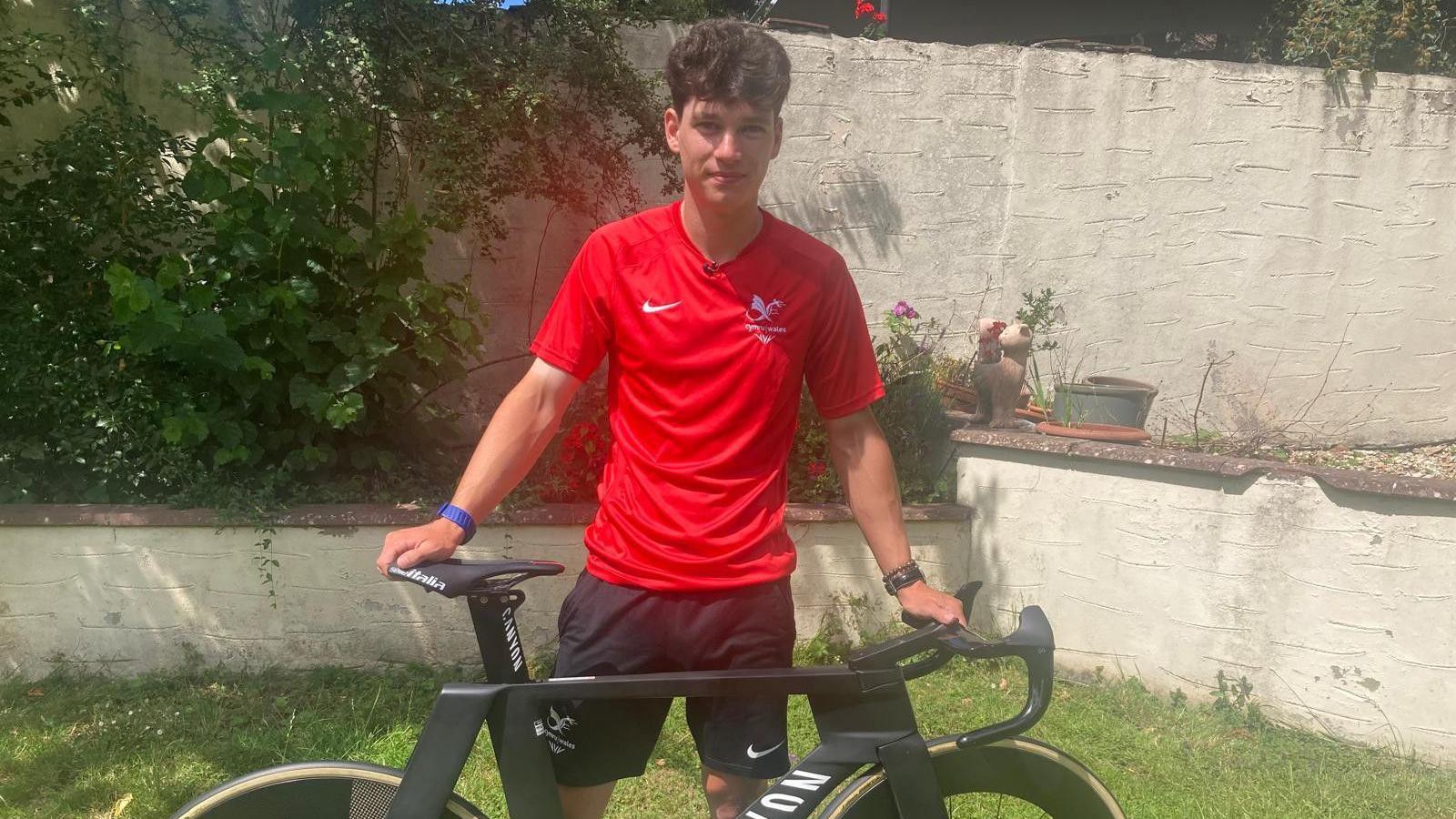 Will Salter standing in a garden with his bike in front of him. He is wearing a red Team Wales Commonwealth Games t-shirt
