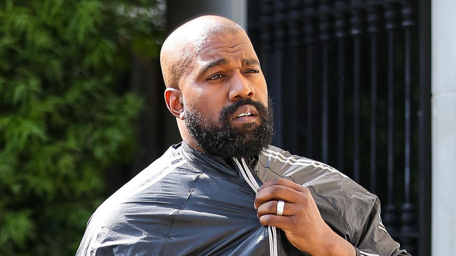 Kanye West in a black jacket. He has a beard and a shaved head. Behind him are some leaves on a tree