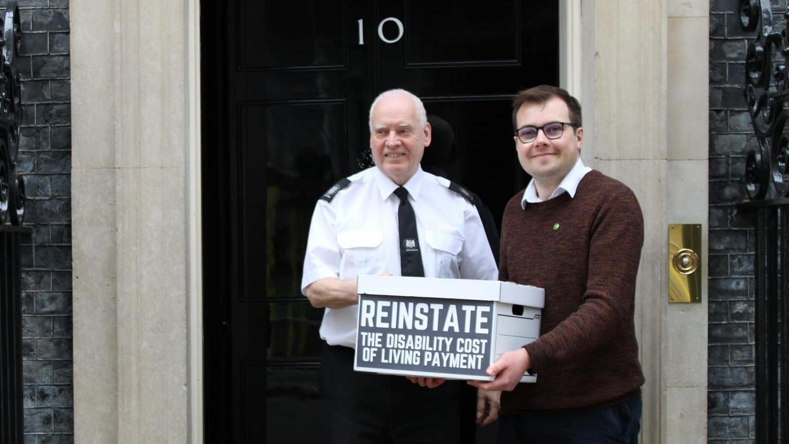 Tom Howard, on the right of the image, wearing a brown shirt over a white shirt while standing outside Downing Street holding a cardboard box with the word REINSTATE on it He is standing next to a security guard.