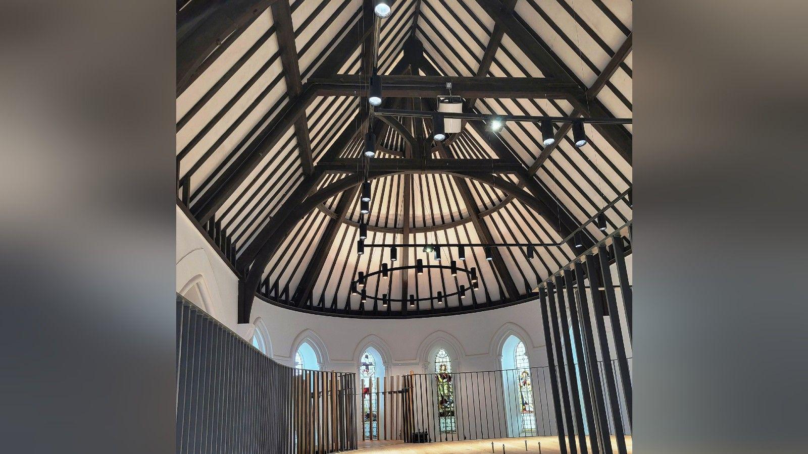 A shot of the inside of Christ Church. The image is taken from a mezzanine level and shows a vaulted ceiling with wooden beams and stained glass windows all around the higher part of the wall. The roof is more like that of a medieval banquet hall than a traditional church, and the lighting has been designed to follow a similar style. The walls are plain white.