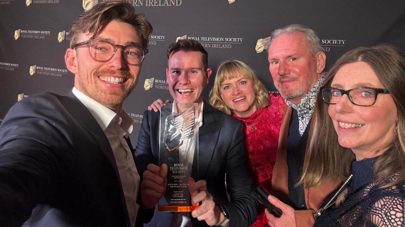 Five people are in a selfie. Three men and two women. They're smiling and one is holding an award.