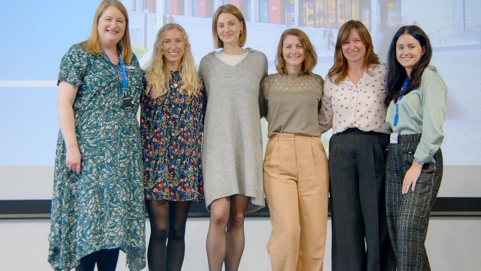 Sofiia Rozhdestvina is pictured with five other women standing in a row smiling at the camera. She is wearing dark patterned trousers and a light green top and has long dark hair.