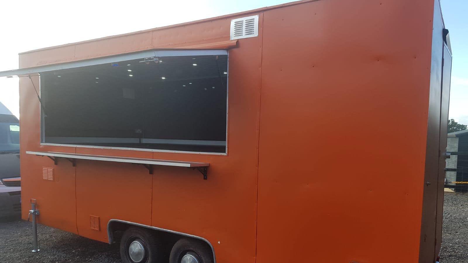 A side view of a large orange trailer parked on some gravel. A serving hatch is open showing ceiling lights inside the trailer. It has wheels in the centre of the trailer