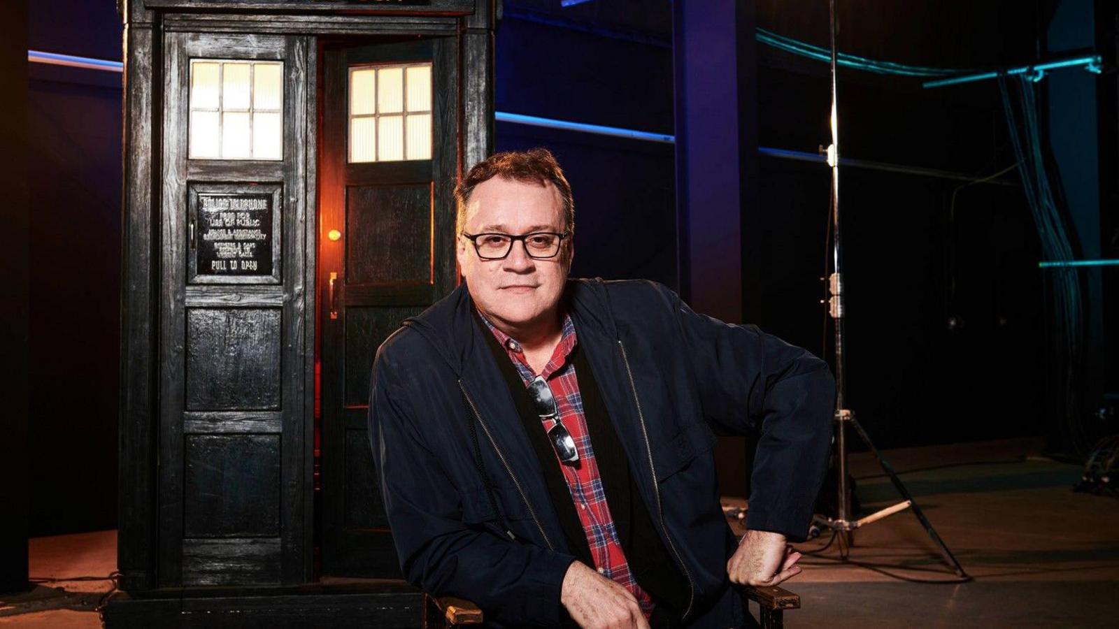 Yn y llun yma mae Russell T Davies yn gwisgo crys coch a glas gyda siaced ddu. Mae'n llun proffesiynol a dynnwyd ar set Dr Who. Gallwch weld y Tardis yn y cefndir. Mae ganddo wallt tywyll ac mae'n gwisgo sbectol ddu.