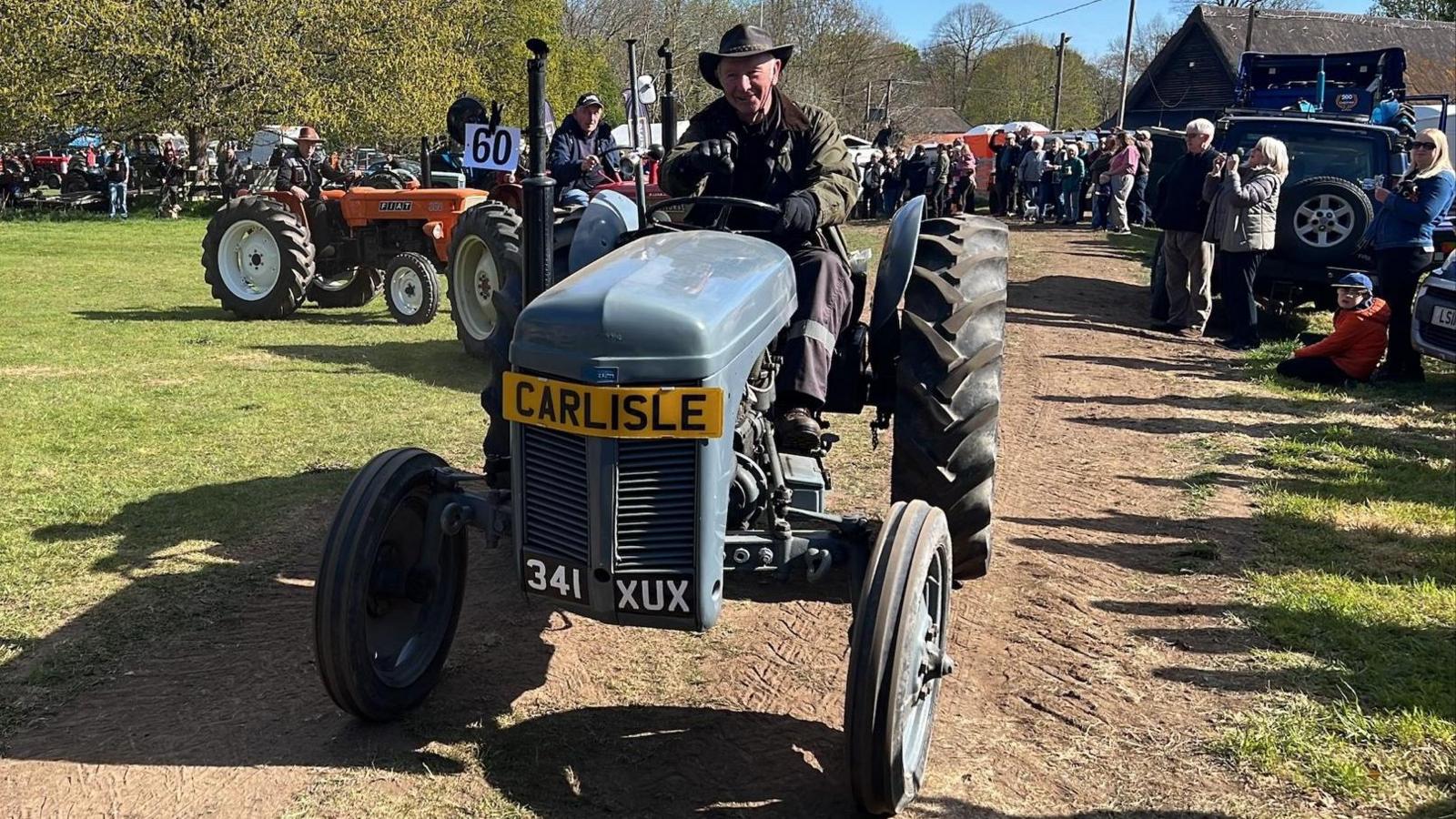 A man wearing a green jacket and hat drives a grey tractor along a dirt track. Behind him are lots of other tractors and people enjoying the event.
