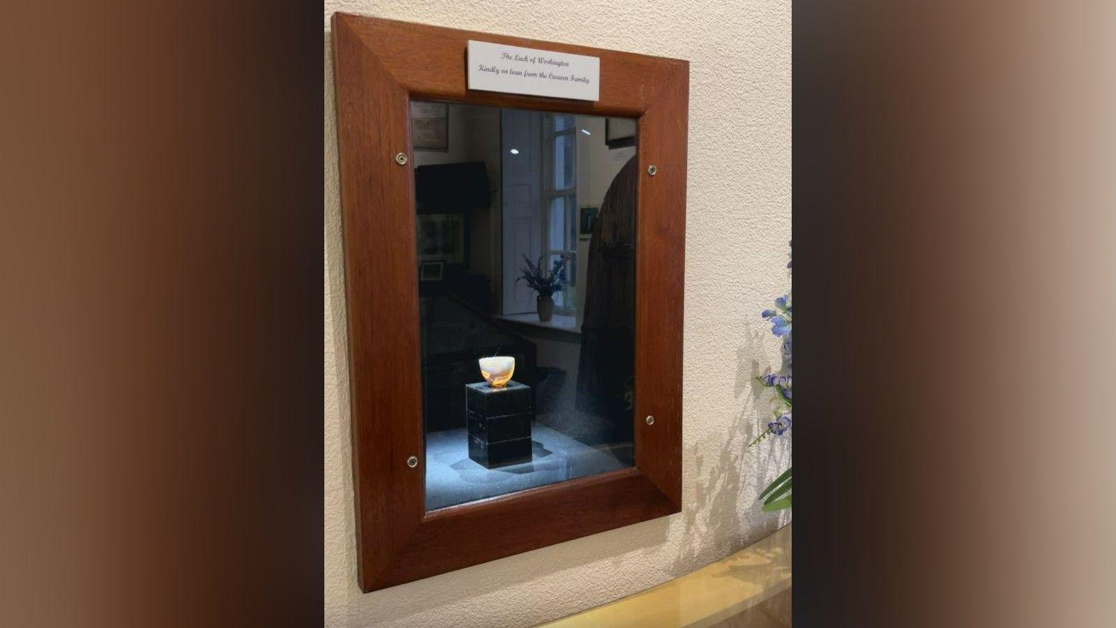 The cup inside a display cabinet set within a wall at the Helena Thompson Museum. The small cup, with a yellow and white swirled motif, is on top of a black plinth. There is a wooden frame around the display cabinet with a silver plaque with its name on the top.