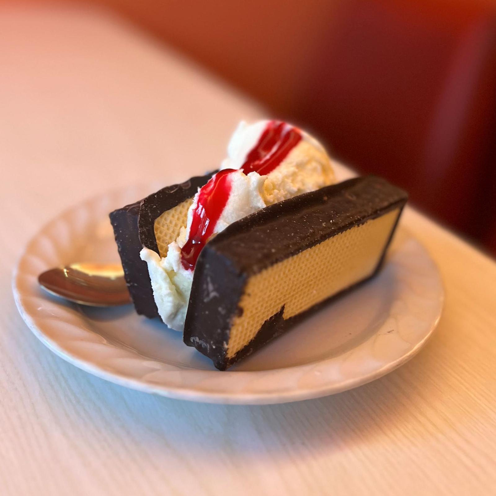 Two scoops of ice cream covered in sauce are sandwiched between chocolate covered, mallow-filled wafers. The treat sits on a white plate on a table.