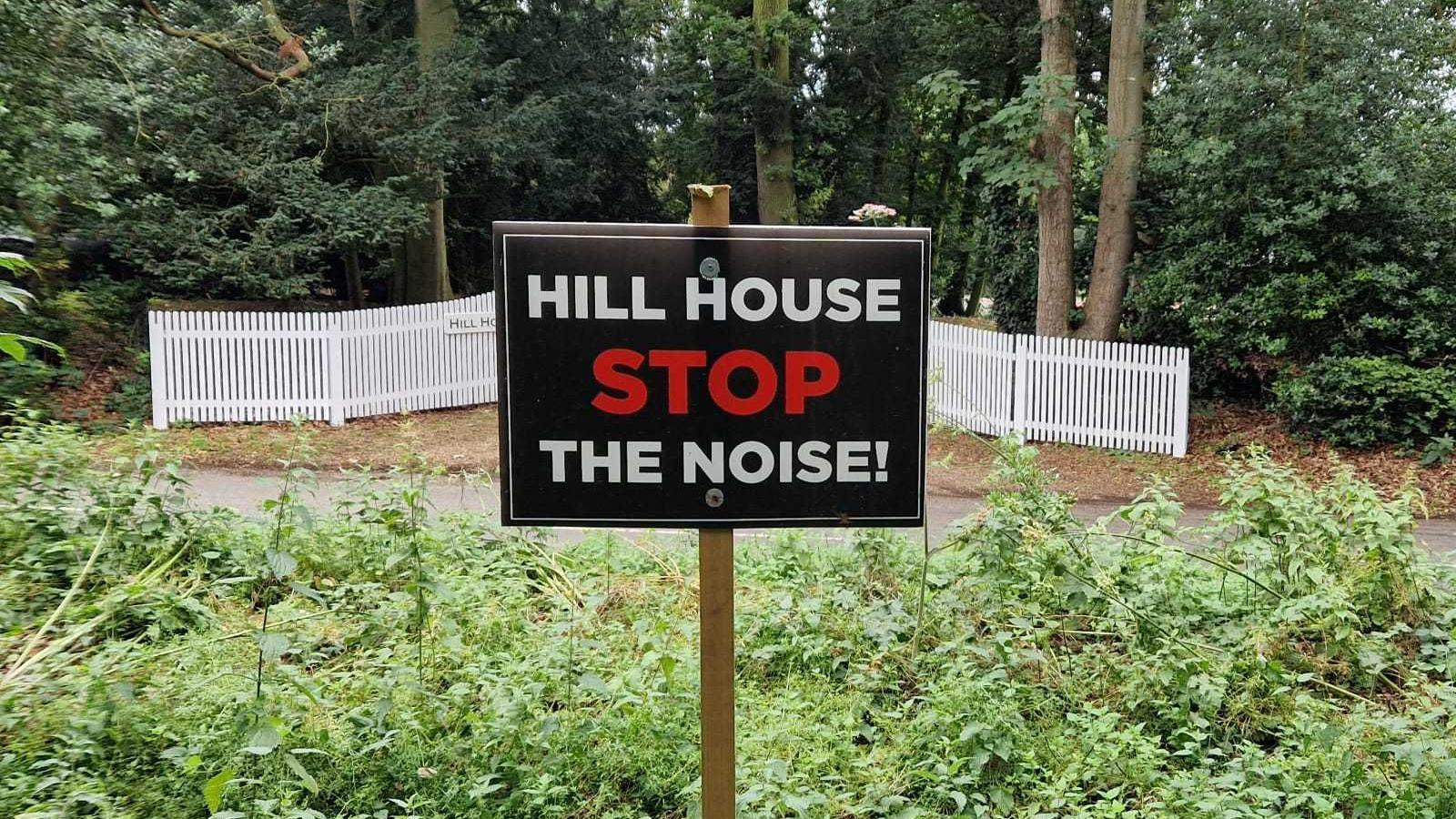 A sign reads: "Hill House. Stop the noise!" The sign has been placed in a bush in front of a road and the entrance to a property. The entrance is lined with white gates.