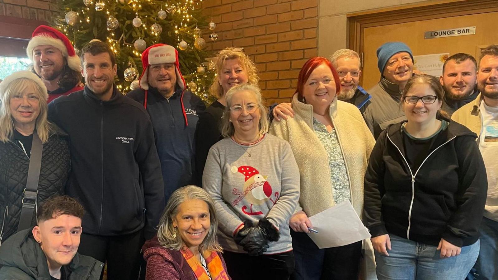 A group of adults huddle together in-front of a Christmas tree. Some wear festive jumpers and santa hats.