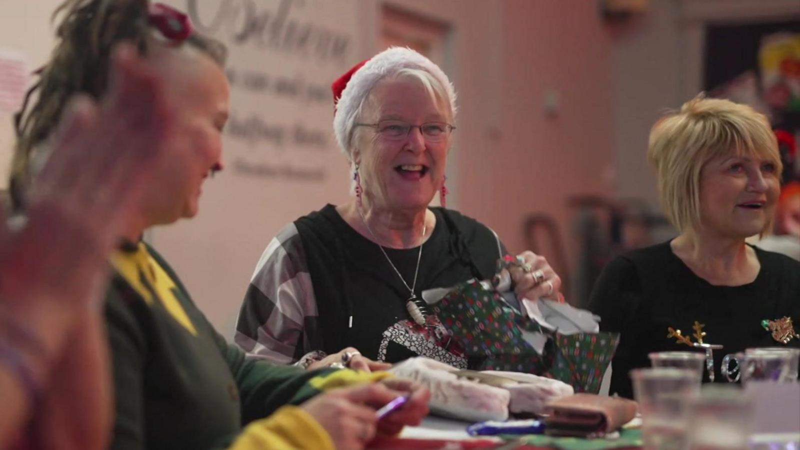 Sandy Withe, wearing a santa hat, laughs as she unwraps a gift while surrounded by other women at a table duing a Christmas party.