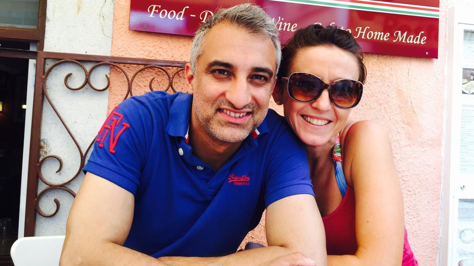 A man and a woman sitting outside, in front of a restaurant. She has dark brown, tied-back hair and is wearing large sunglasses and a pink top. The man has short grey hair and is wearing a blue, short-sleeved top with red lettering on it.